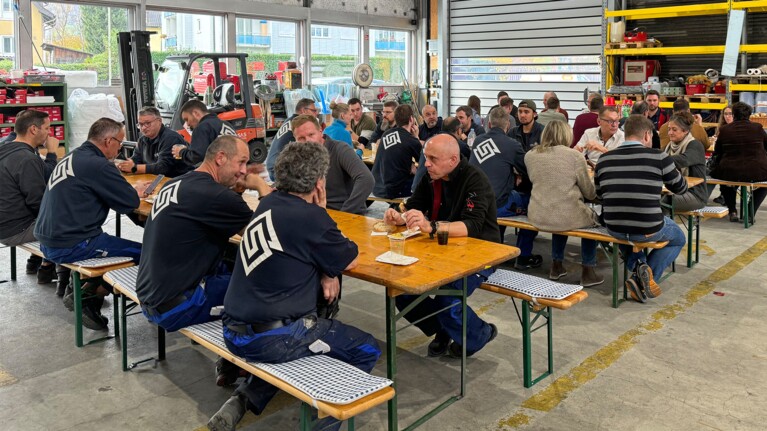 Die Belegschaft von Surber Metallbau sitzt an mehreren Bierbänken verteilt in der Produktionshalle und isst Bratwurst.