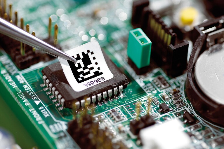 Une étiquette est collée sur un circuit imprimé à l'aide d'une pincette.